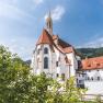 Historisches Gebäude mit rotem Dach und Turm, umgeben von Bäumen.