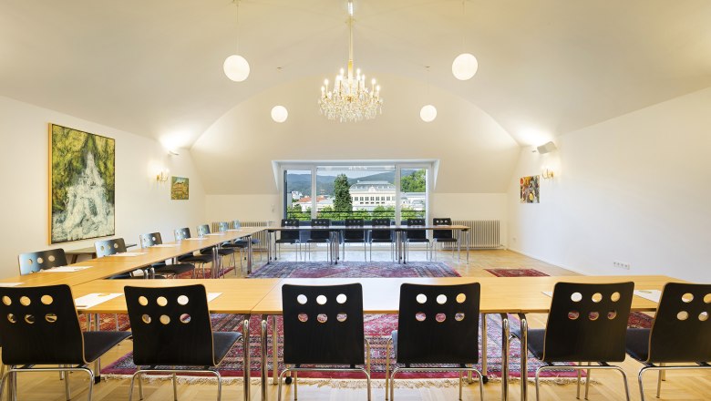 Ein Konferenzraum mit U-förmiger Tischanordnung, Stühlen, einem Kronleuchter und großen Fenstern mit Blick auf ein Gebäude und Berge.