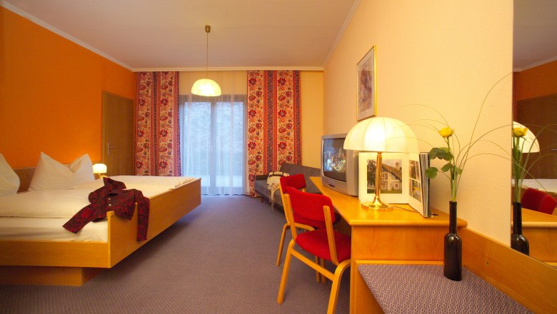 Hotelzimmer mit Doppelbett, rotem Stuhl, Fernseher und Lampe auf einem Tisch. Gelbe Wand und Vorh&auml;nge mit Blumenmuster.