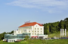 Landzeit Steinhäusl Gebäude mit rotem Dach und Glasfassade, umgeben von Bäumen und blauen Himmel.