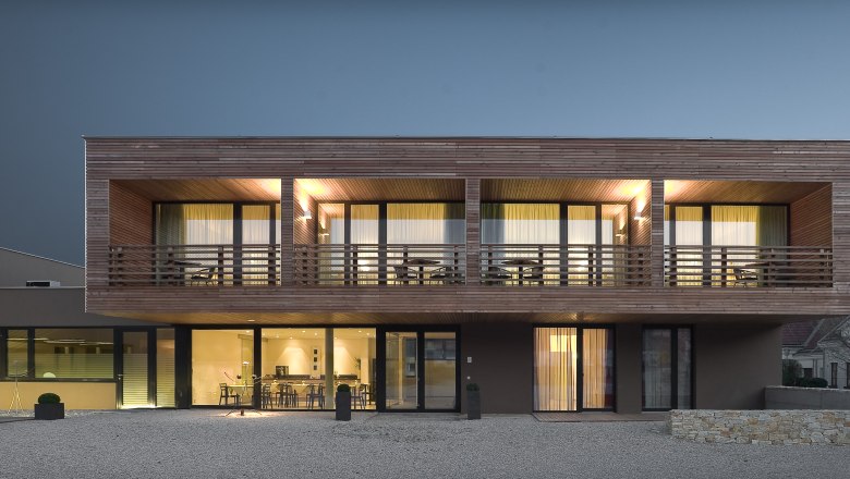 Modernes zweist&ouml;ckiges Haus mit Holzverkleidung und gro&szlig;en Fenstern bei Nachtbeleuchtung.
