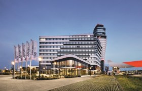 Modernes Geb&auml;ude am Flughafen Wien bei D&auml;mmerung mit Flaggen und Kontrollturm im Hintergrund.
