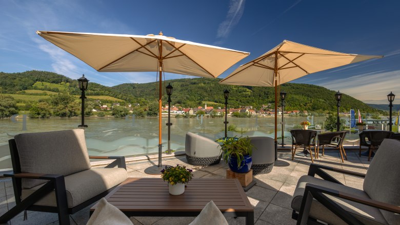 Terrasse mit Blick auf die Donau und umliegende H&uuml;gel, ausgestattet mit Sonnenschirmen und Sitzm&ouml;beln.