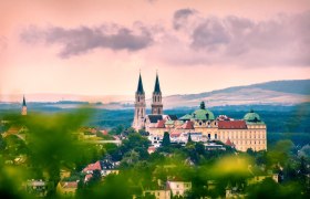 Panoramablick auf das Stift Klosterneuburg mit zwei Kirchtürmen und barockem Gebäude, umgeben von grüner Landschaft und unter bewölktem Himmel.