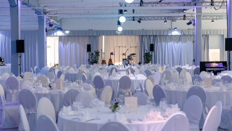 Eleganter Veranstaltungsraum mit runden, weiß gedeckten Tischen und Bühne mit Musikinstrumenten.