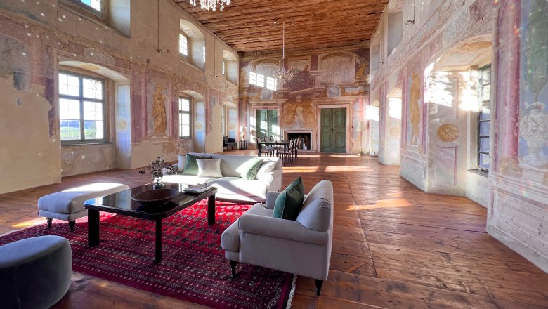 Ein historischer Festsaal mit Holzboden, bemalten W&auml;nden und einem Kronleuchter. Moderne M&ouml;bel und ein roter Teppich sind im Raum platziert.