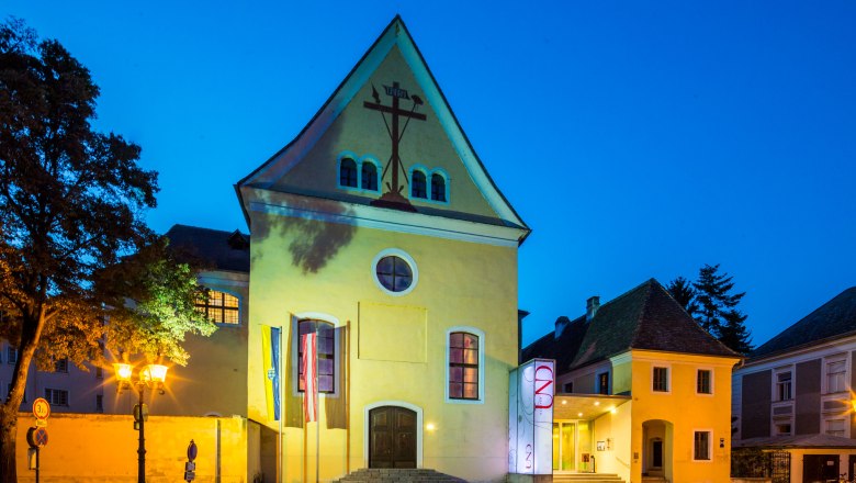 Gelbes Gebäude mit Kreuzsymbol, beleuchtet bei Nacht.