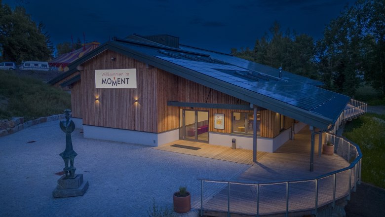 Ein modernes Geb&auml;ude mit Holzverkleidung und Solarpanelen auf dem Dach, beleuchtet in der Abendd&auml;mmerung. Ein Schild mit der Aufschrift 'Willkommen im MOMENT' ist sichtbar.