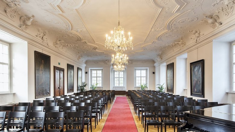 Eleganter Saal mit Stuckdecke, Kronleuchtern, Stuhlreihen und rotem Teppich.