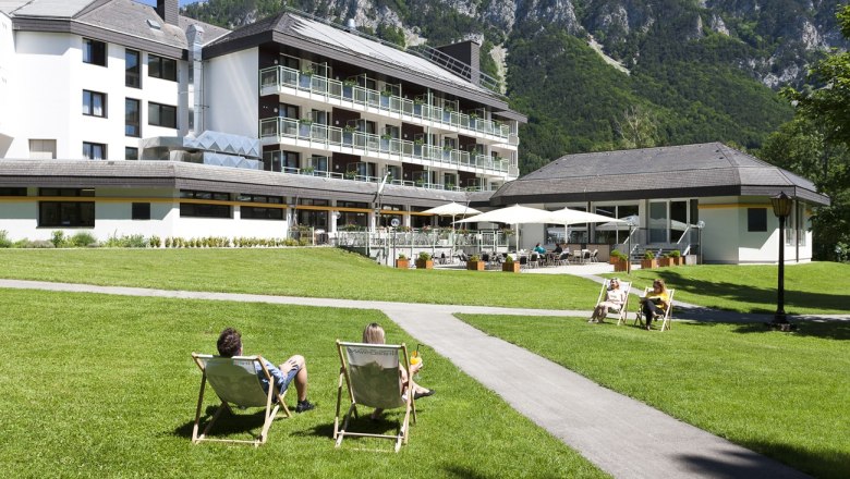 Ein Hotel mit Terrasse und Liegest&uuml;hlen auf einer Wiese, umgeben von Bergen.