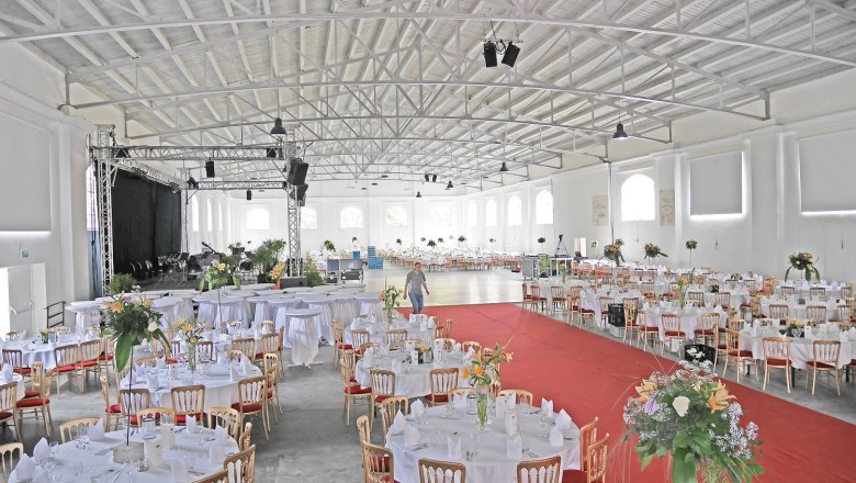 Innenansicht einer großen Halle mit Galabestuhlung, runden Tischen und einer Bühne.
