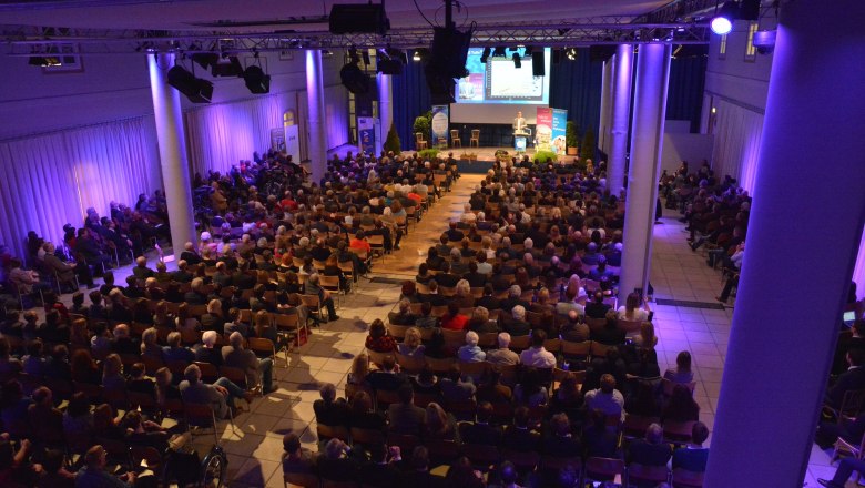 Ein vollbesetzter Veranstaltungsraum mit Bühne und Publikum, beleuchtet in blauem Licht.