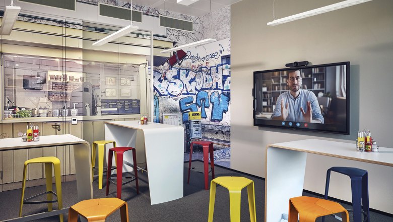 Moderner Konferenzraum mit Graffiti-Wand, bunten Hockern und Videokonferenz-Bildschirm.