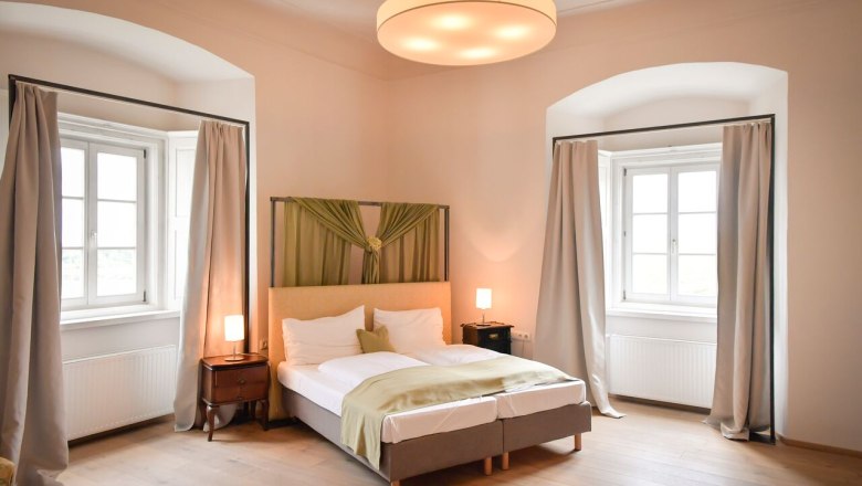 Ein elegantes Schlafzimmer mit Doppelbett, zwei Fenstern mit Vorh&auml;ngen und stilvoller Beleuchtung.