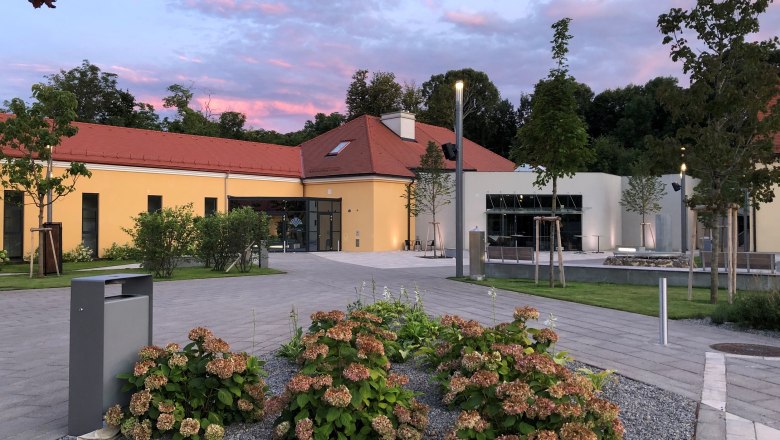 Modernes Gebäude mit gelber Fassade und rotem Dach, umgeben von Pflanzen und Bäumen, bei Sonnenuntergang.