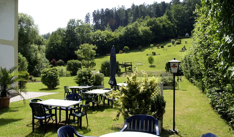 Garten mit Tischen und St&uuml;hlen, umgeben von B&auml;umen und Wiesen.