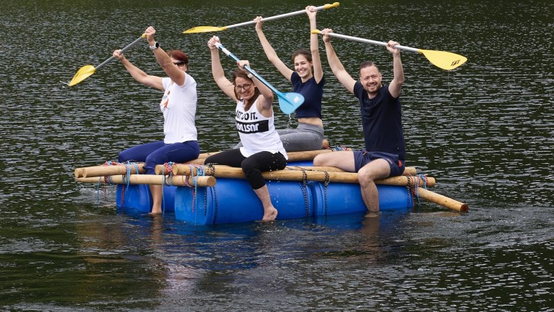 Vier Personen sitzen auf einem selbstgebauten Flo&szlig; aus blauen F&auml;ssern und Holzstangen auf einem See und heben fr&ouml;hlich ihre Paddel in die Luft.