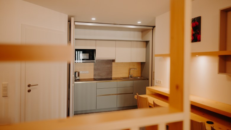 Moderne K&uuml;chenzeile in einem Apartment in hellen Farbt&ouml;nen. Man sieht eine Sp&uuml;le, eine Mikrowelle, eine gro&szlig;e Herdplatte und einen Wasserkocher. Die K&uuml;che kann hinter einer verschiebbaren Trennwand versteckt werden. Vor der K&uuml;che sieht man einen Tisch an der Wand und 4 Sessel.