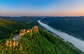Luftaufnahme der Burgruine Aggstein an der Donau, umgeben von grünen Hügeln und Wäldern bei Sonnenuntergang.