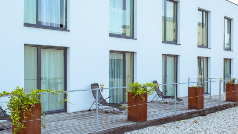 Moderne Terrasse mit Liegest&uuml;hlen und Pflanzen vor einem Geb&auml;ude mit gro&szlig;en Fenstern.
