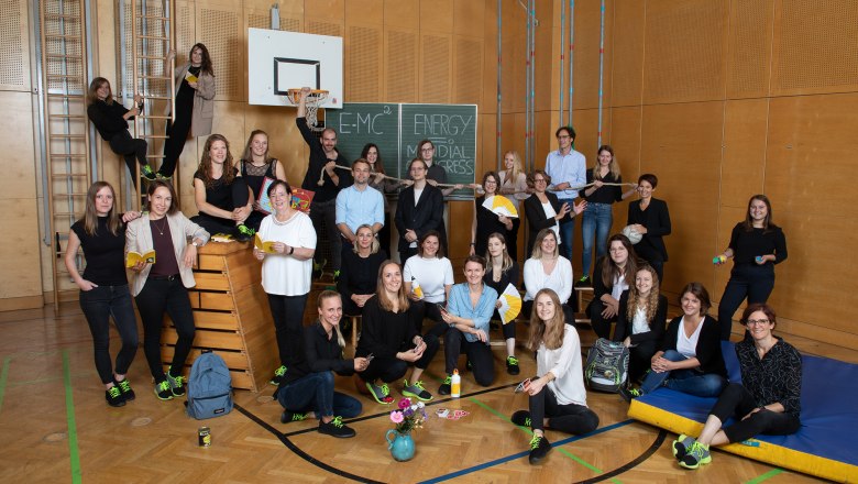 Gruppe von Menschen in einer Turnhalle mit Sportgeräten und Tafel im Hintergrund.