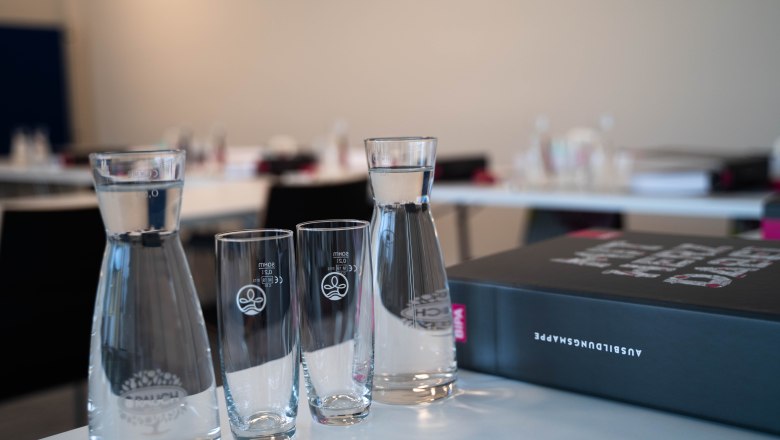 Ein Seminarraum mit Wasserkaraffen, Gläsern und einem Ordner auf einem Tisch.