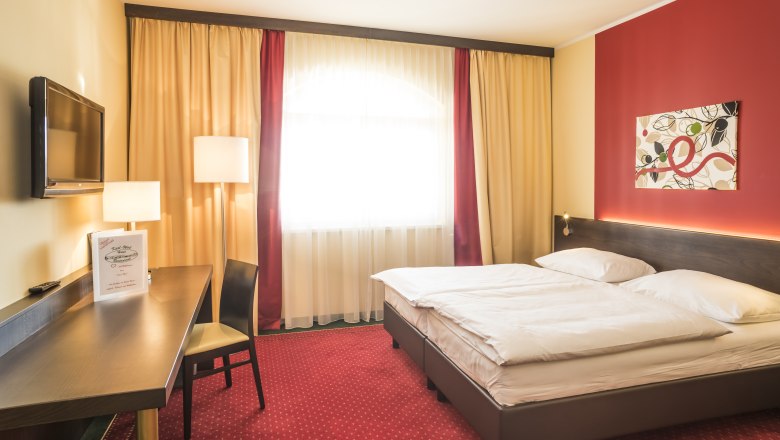 Ein gem&uuml;tliches Hotelzimmer mit Doppelbett, rotem Teppich, Schreibtisch, Fernseher und moderner Kunst an der Wand.