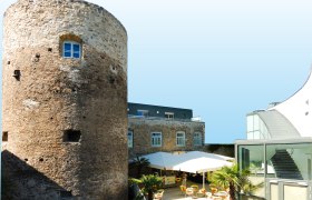 Historischer Turm neben modernem Geb&auml;ude mit Terrasse und Sonnenschirmen.