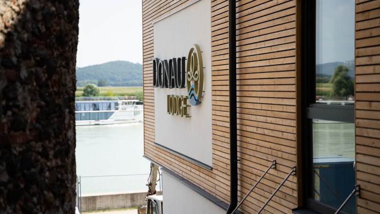 Gebäude mit Holzverkleidung und Schriftzug 'Donau Lodge', im Hintergrund die Donau und ein Schiff.