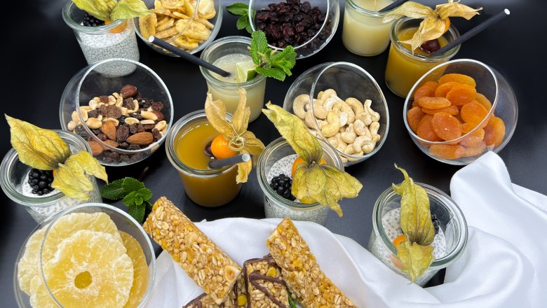 Eine Auswahl an Snacks und Getr&auml;nken, darunter N&uuml;sse, Trockenfr&uuml;chte, Chia-Pudding und S&auml;fte, auf einem Tisch arrangiert.