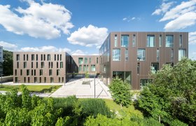 Moderne Universitätsgebäude mit brauner Fassade und großen Fenstern, umgeben von grüner Vegetation unter blauem Himmel.